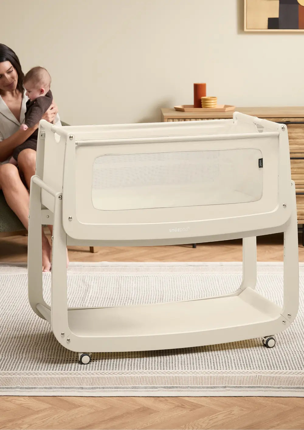 Das freistehende, beige Snüz Beistellbett SnüzPod 5 Cashmere mit praktischen Rollen am Gestell und atmungsaktivem Stoffbezug; im Hintergrund sitzt eine Mutter mit Kind.