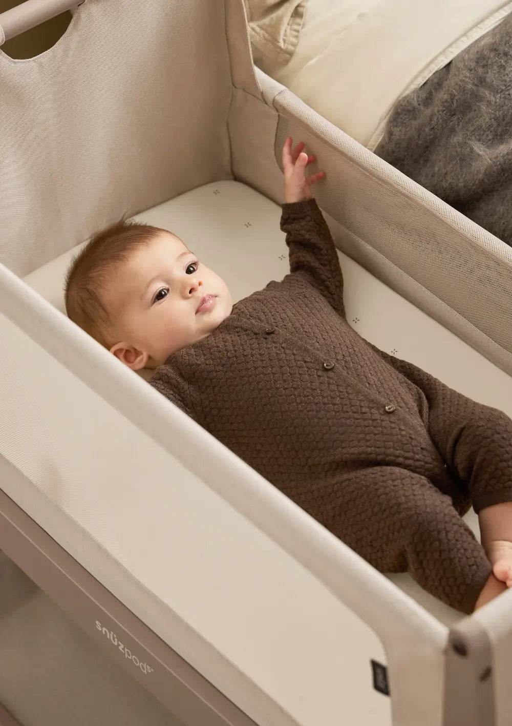 Ein Baby im braunen Strampler liegt entspannt auf der Matratze des Snüz Beistellbettes SnüzPod 5 Mocha und streckt eine Hand nach oben.