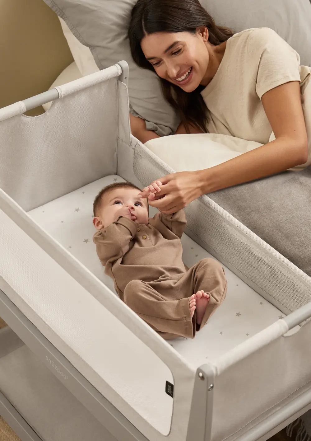 Eine Mutter liegt lächelnd im großen Bett direkt neben dem geöffneten grauen Snüz Beistellbett SnüzPod 5 Pebble und hält die Hand ihres Babys; sicheres Co-Sleeping.
