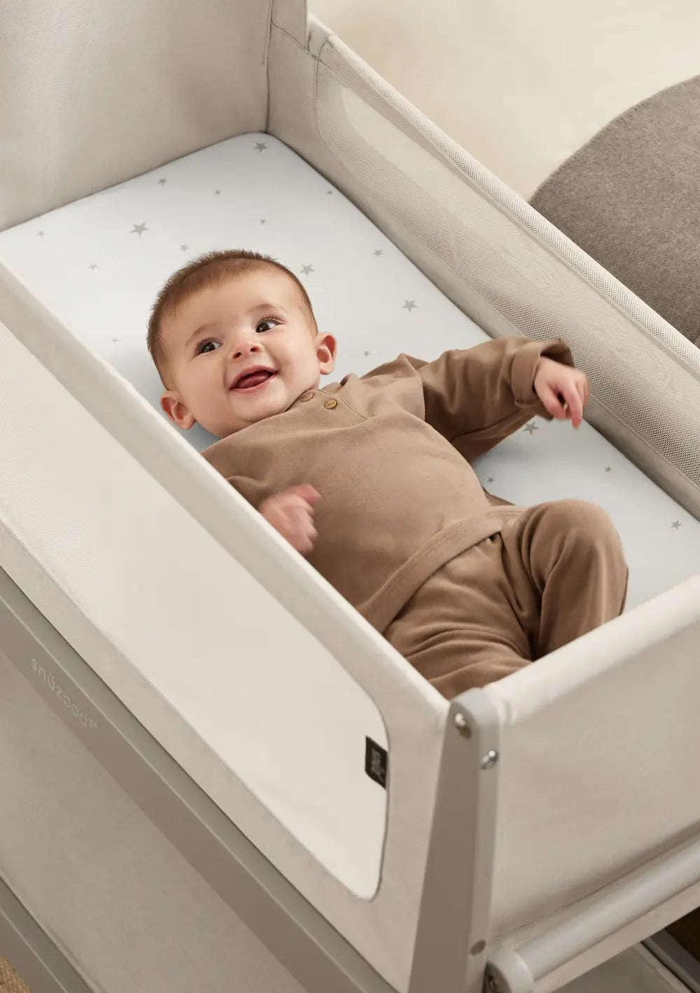 Ein fröhliches Baby liegt auf einer Matratze mit Sternenmuster im grauen Snüz Beistellbett SnüzPod 5 Pebble; halbe Draufsicht auf den geschützten Schlafbereich.