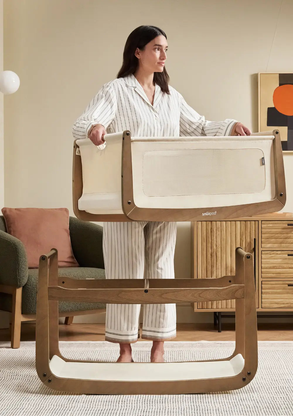 Eine Frau demonstriert das praktische Design des Snüz Beistellbettes SnüzPod 5 Walnuss, indem sie die obere Liegewanne vom walnussfarbenen Holzuntergestell abhebt.