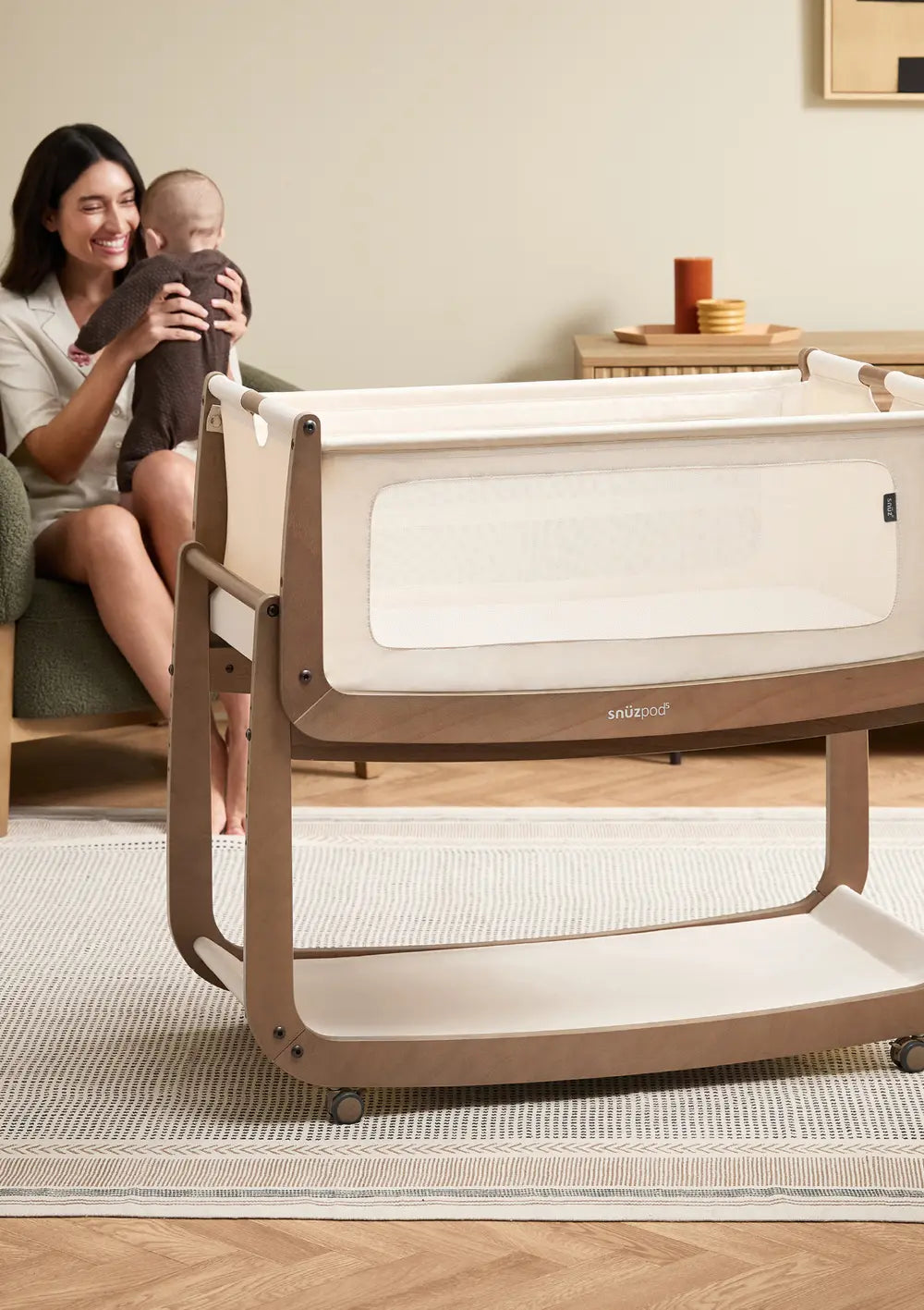 Das freistehende Snüz Beistellbett SnüzPod 5 Walnuss mit montierten Rollen im Schlafzimmer; im Hintergrund sitzt die Mutter mit dem Baby auf dem Schoss.