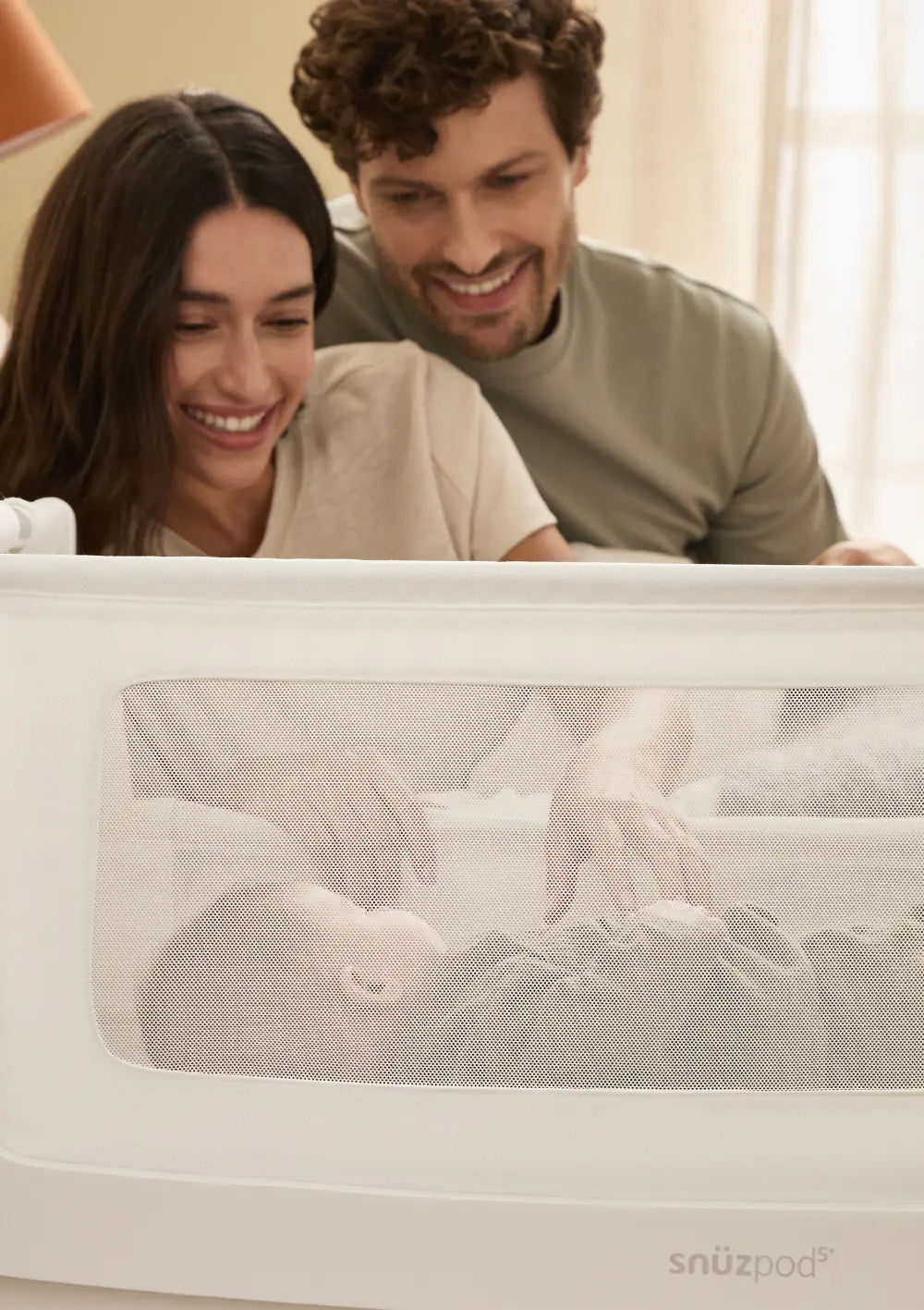 Ein glückliches Elternpaar blickt von oben durch das atmungsaktive Mesh-Sichtfenster des weißen Snüz Beistellbettes SnüzPod 5 auf das schlafende Baby.