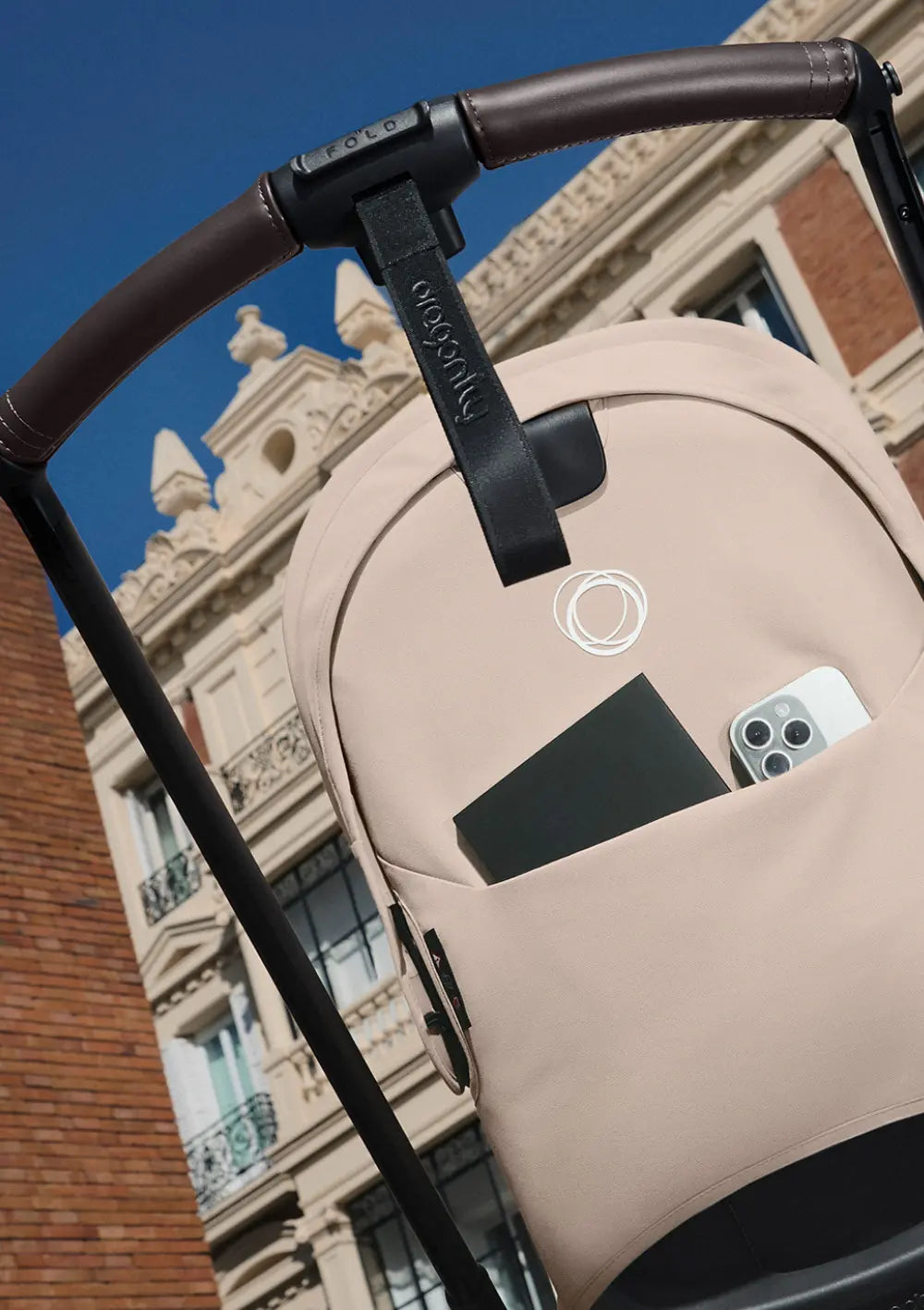 Funktionale Detailansicht der praktischen Aufbewahrungstasche an der Rückenlehne des Bugaboo Kinderwagens Dragonfly Plus, gefüllt mit Smartphone und Geldbörse, direkt unter dem braunen Leder-Schiebebügel.