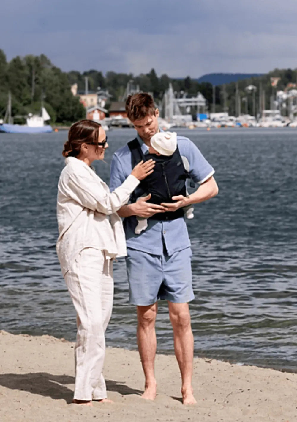 Eine Familie am Strand: Die Mutter berührt zärtlich das schlafende Baby, das der Vater in der schwarzen Voksi Babytrage Huddle Black vor dem Bauch trägt; maritimes Hafen-Panorama im Hintergrund.