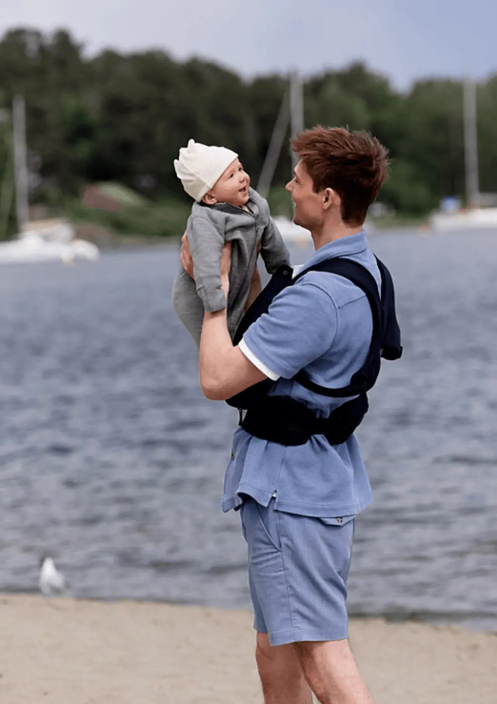 Ein Mann im blauen Outfit steht am Sandstrand, trägt die Voksi Babytrage Huddle Black einsatzbereit am Körper und hält sein glückliches Baby lächelnd in die Luft.