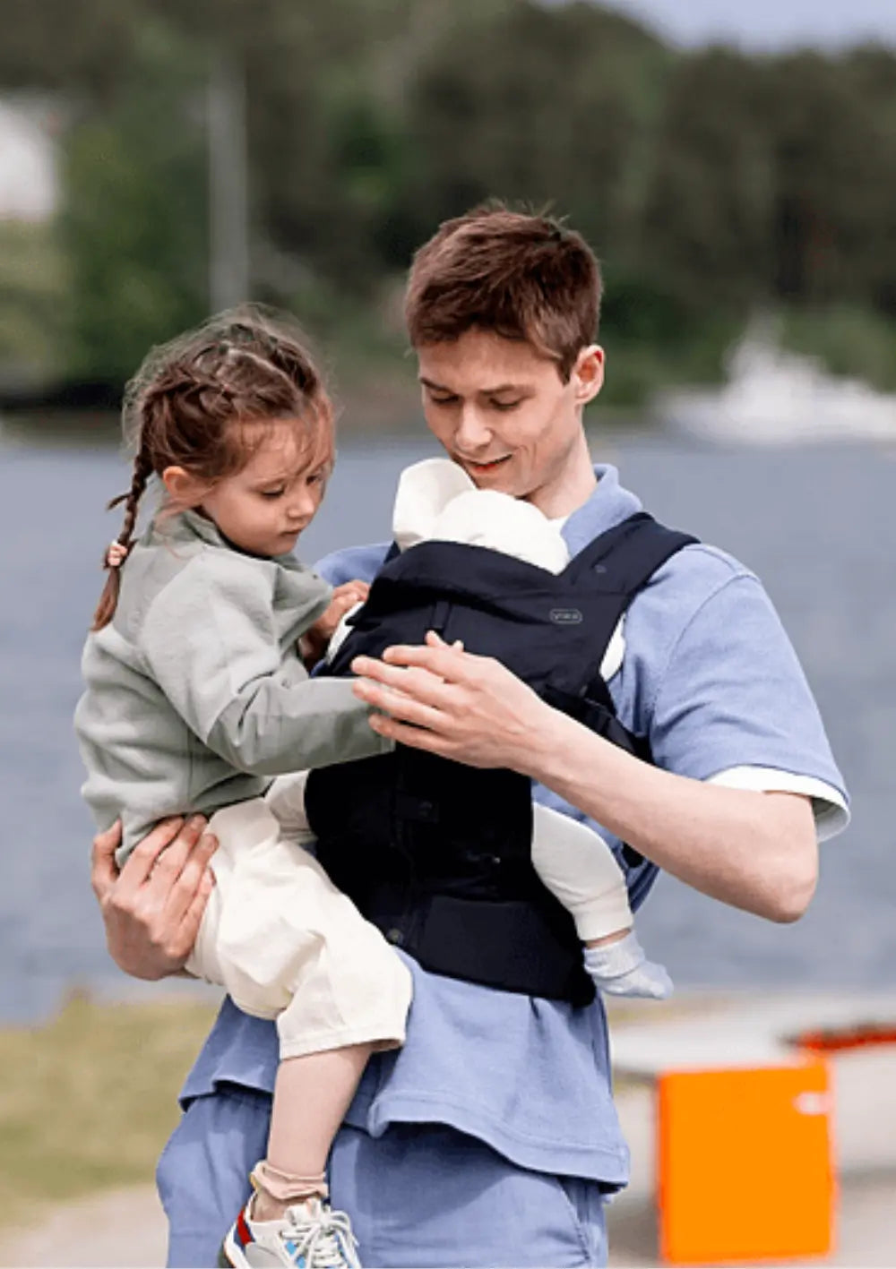 Ein Vater hält seine Tochter im Arm, während sein Baby sicher und geborgen in der ergonomischen Voksi Babytrage Huddle Black an seiner Brust positioniert ist; im Hintergrund eine Wasserfläche.