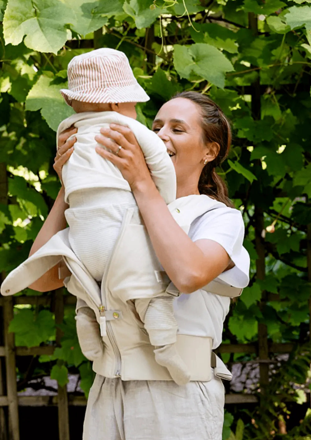 Eine glückliche Mutter hebt ihr Baby beim Spaziergang leicht an, um das einfache Handling und den bequemen Sitz der hellen Voksi Babytrage Huddle Cream zu zeigen.