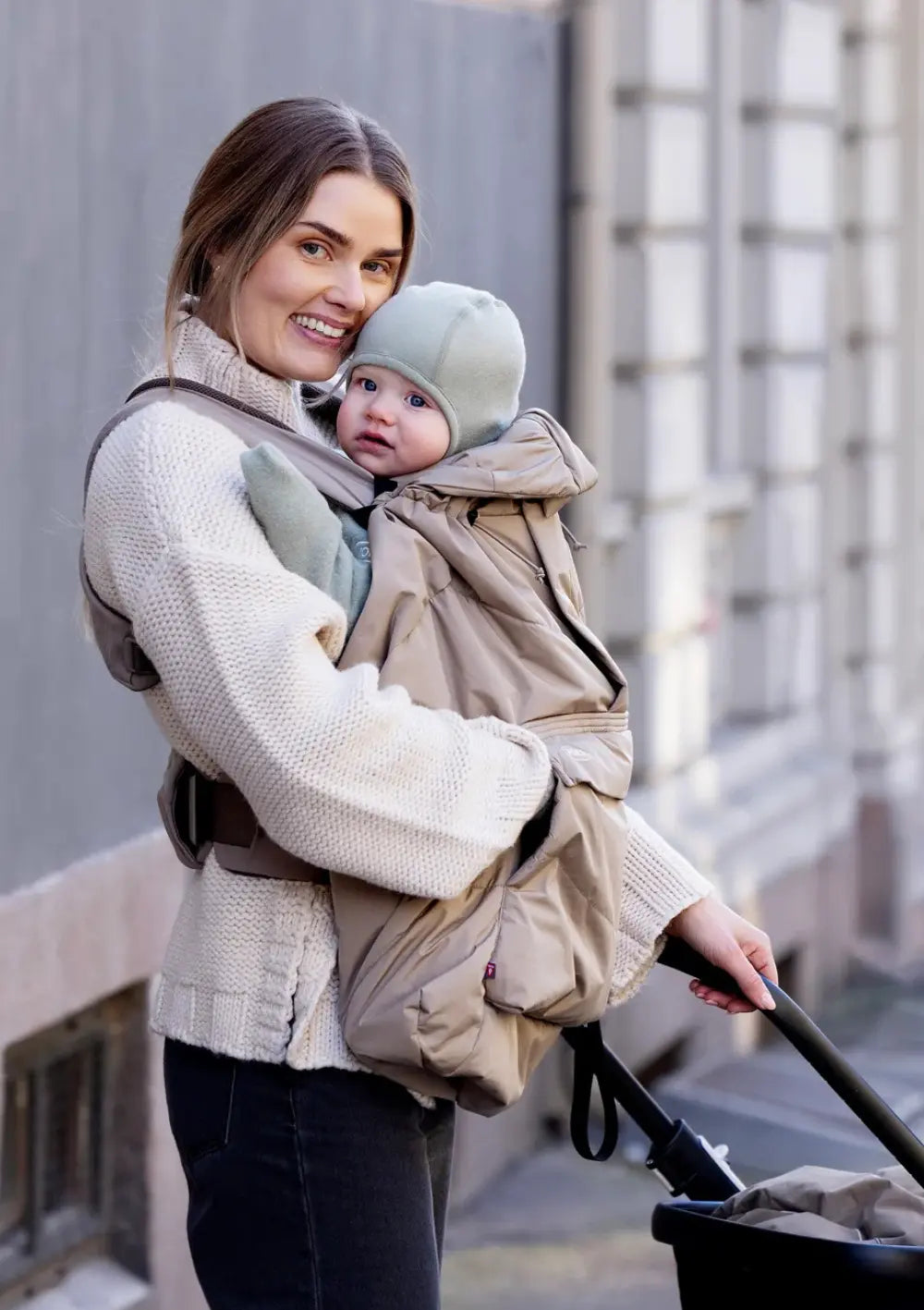 Porträtaufnahme einer Mutter mit Schirmmütze und ihrem Baby, das durch das gesteppte Voksi Cover für Babytragen Dark Sand mit praktischer Kapuze warm eingepackt ist.