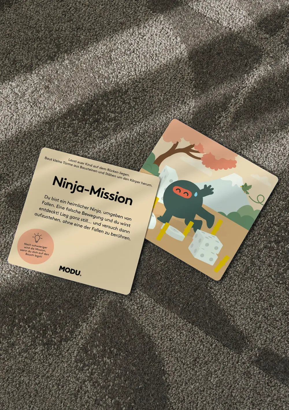 Zwei quadratische Karten aus dem MODU Spielkarten-Set liegen nebeneinander auf dem Teppich; eine zeigt die Spielanleitung für die „Ninja-Mission“, die andere eine passende Comic-Illustration.