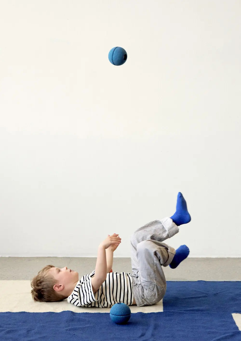 Ein Kind liegt auf einer Spielmatte, hat die Beine hochgestreckt und wirft einen dunkelblauen Ball aus dem MODU 3er-Set Schaumstoffbälle Blue in die Luft, während ein weiterer Ball neben ihm auf dem Boden liegt.