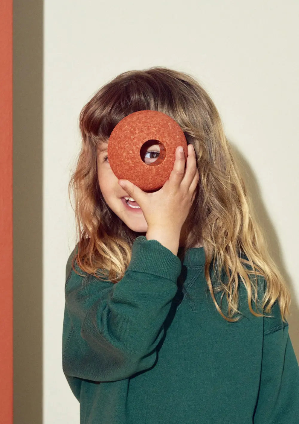 Ein fröhliches Kind blickt spielerisch durch die runde Verbindungsöffnung eines Balles aus dem MODU 3er-Set Schaumstoffbälle Orange, den es sich vor das Auge hält.