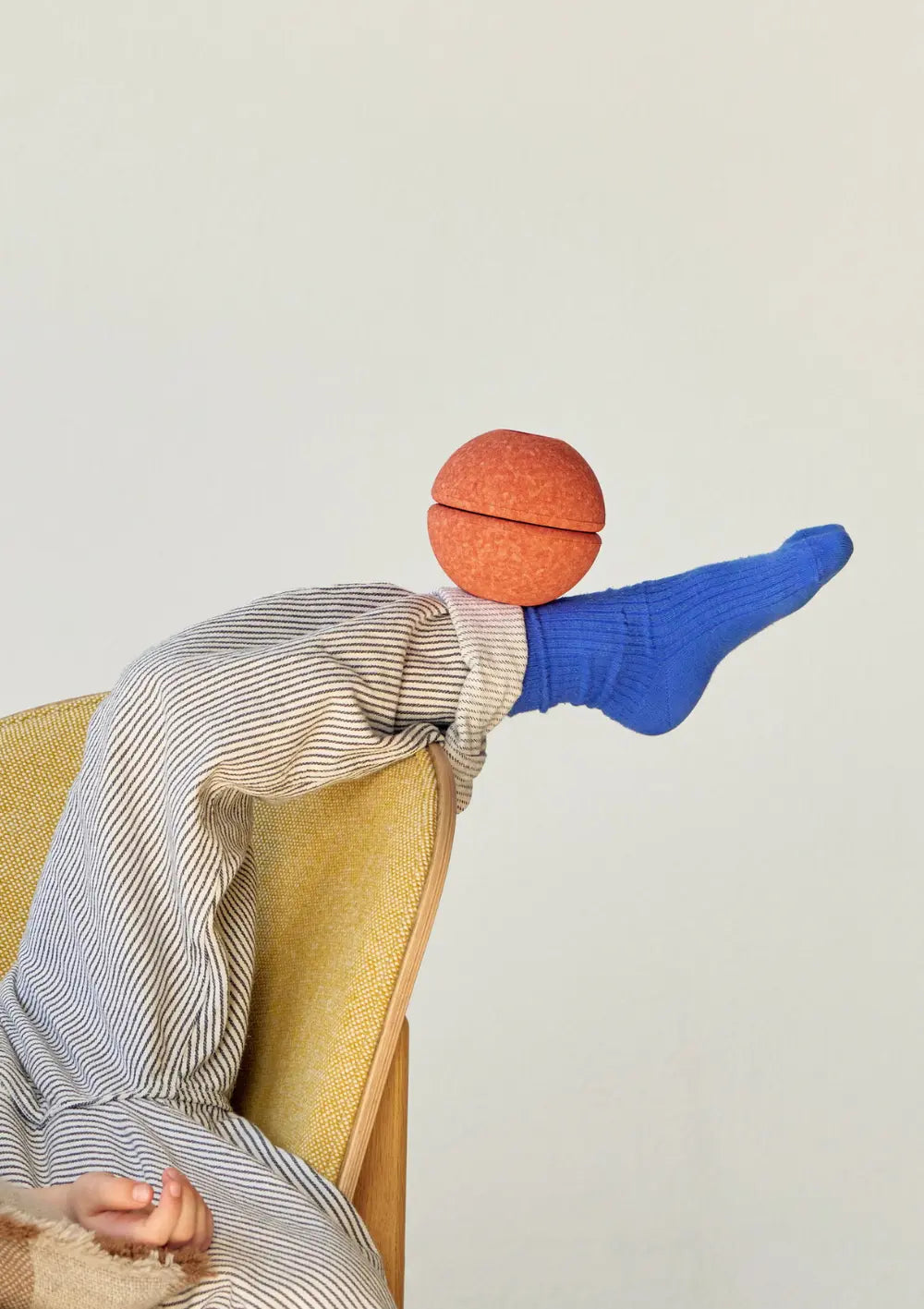 Ein Kind liegt in einem Sessel und balanciert mit hochgestrecktem Bein einen runden Ball aus dem MODU 3er-Set Schaumstoffbälle Orange geschickt auf seinem Fuß mit blauer Socke.