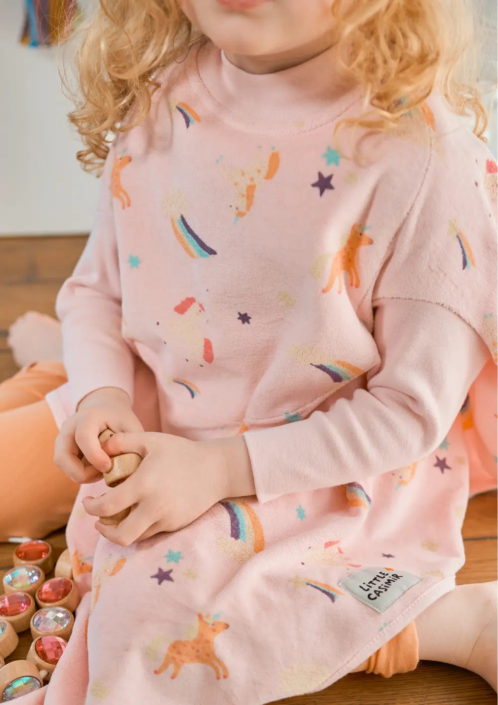 Nahaufnahme eines sitzenden Kindes auf dem Holzboden, das das weiche, rosa Little Casimir Langarm-Lätzchen Boo Rainbow Dreams mit bunten Einhorn- und Sternenmotiven sowie einem aufgenähten Marken-Label trägt.