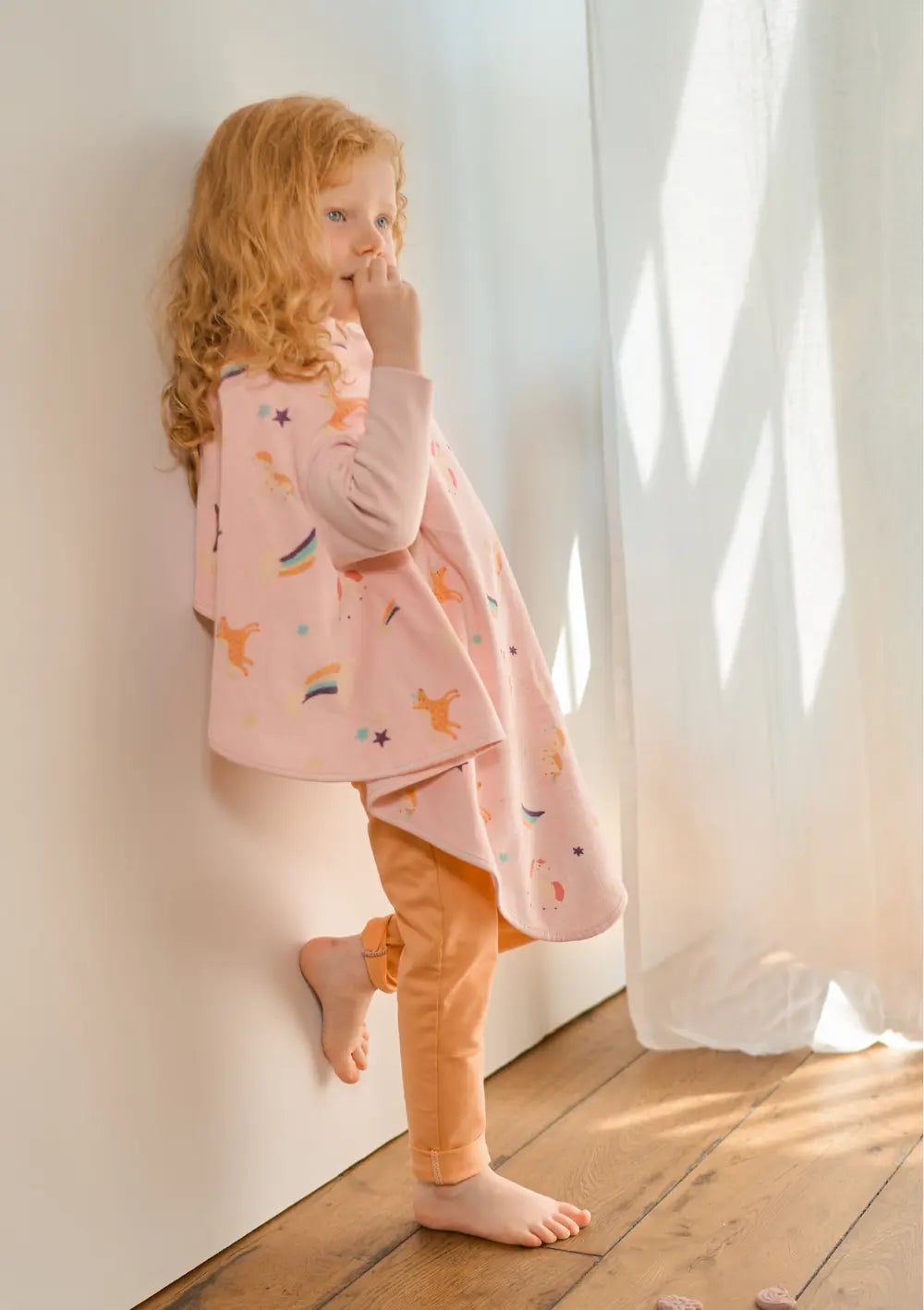 Seitenansicht eines Mädchens, das barfuß an einer hellen Wand steht und das rosa Little Casimir Langarm-Lätzchen Boo Rainbow Dreams trägt; das Frottee-Lätzchen ist mit kleinen Einhörnern und Regenbögen bedruckt.