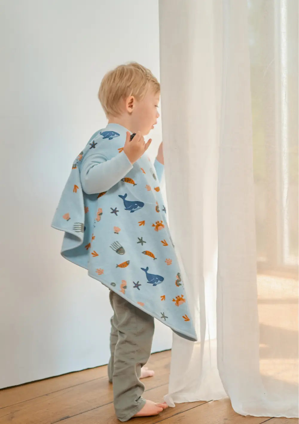 Seitenansicht eines stehenden Kindes im hellblauen Little Casimir Langarm-Lätzchen Boo Deep Sea mit Wal- und Fisch-Motiven; der großzügige Schnitt mit langen Ärmeln bietet optimalen Kleidungsschutz im Alltag.