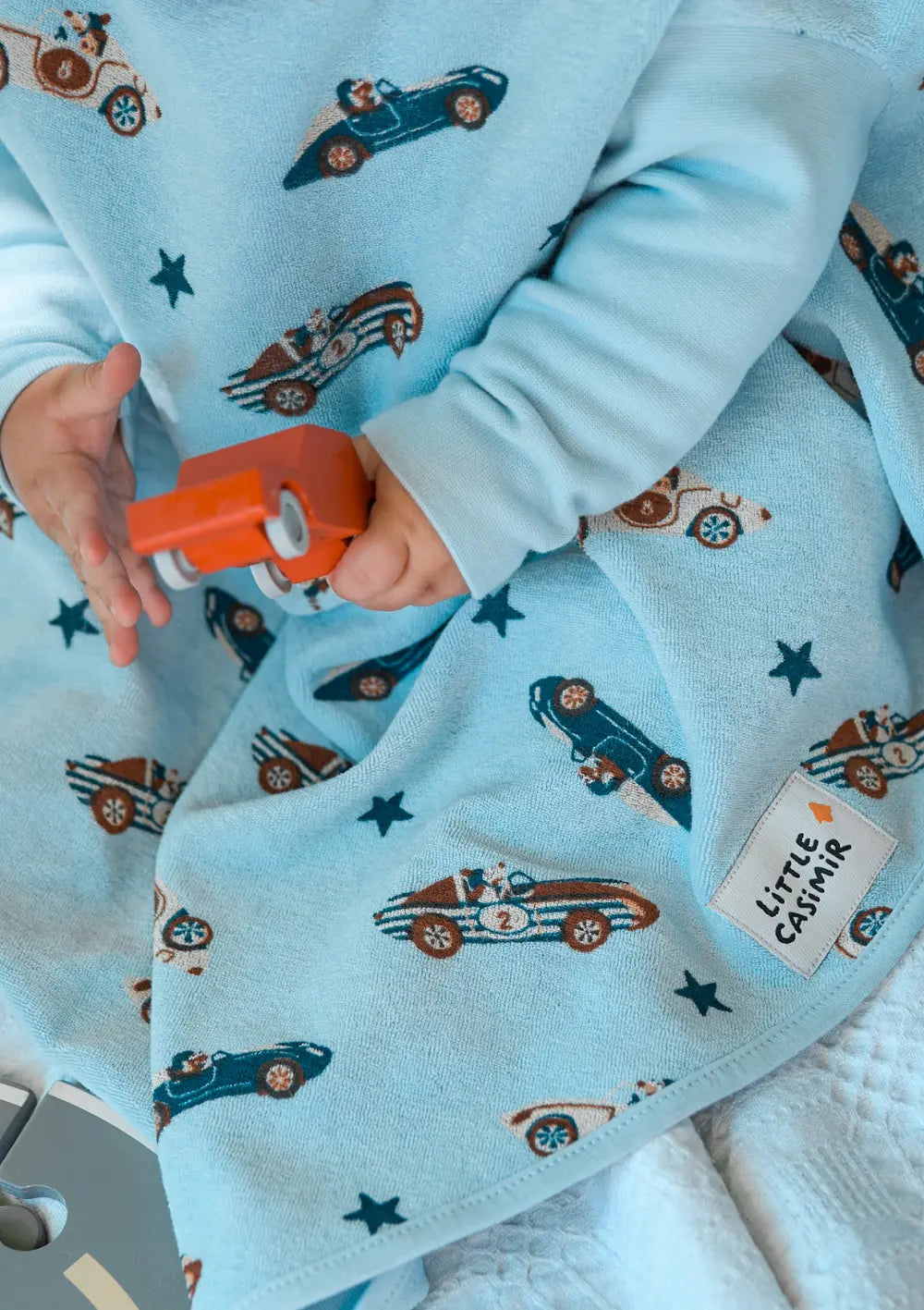 Nahaufnahme eines Kindes, das ein orangefarbenes Spielzeugauto hält; das hellblaue Little Casimir Langarm-Lätzchen Boo Sky Racers aus weichem Frottee zeigt ein Muster aus Rennwagen und kleinen Sternen sowie das Marken-Label.