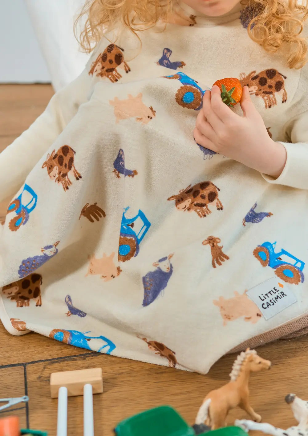 Nahaufnahme eines Kindes auf dem Holzboden, das eine Erdbeere hält; das cremefarbene Little Casimir Langarm-Lätzchen Boo Farm Friends zeigt bunte Motive von Traktoren, Kühen und Schafen.