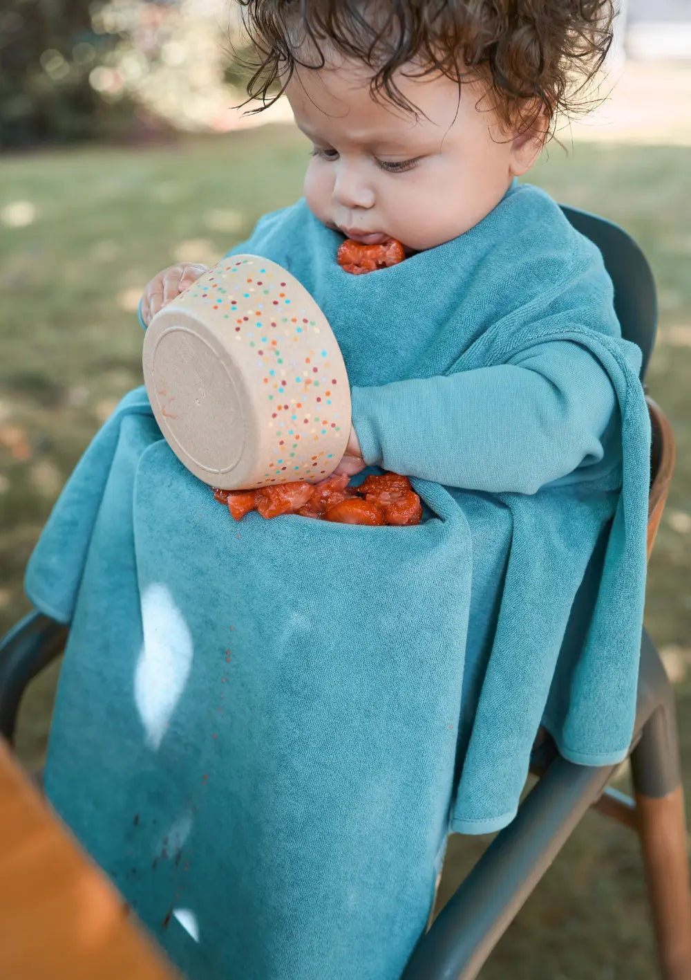 Ein Kleinkind sitzt im Garten im Hochstuhl und isst Erdbeeren; das schützende Little Casimir Langarm-Lätzchen Boo Dusty Ocean fängt herabfallende Fruchtstücke im Schoß zuverlässig auf.