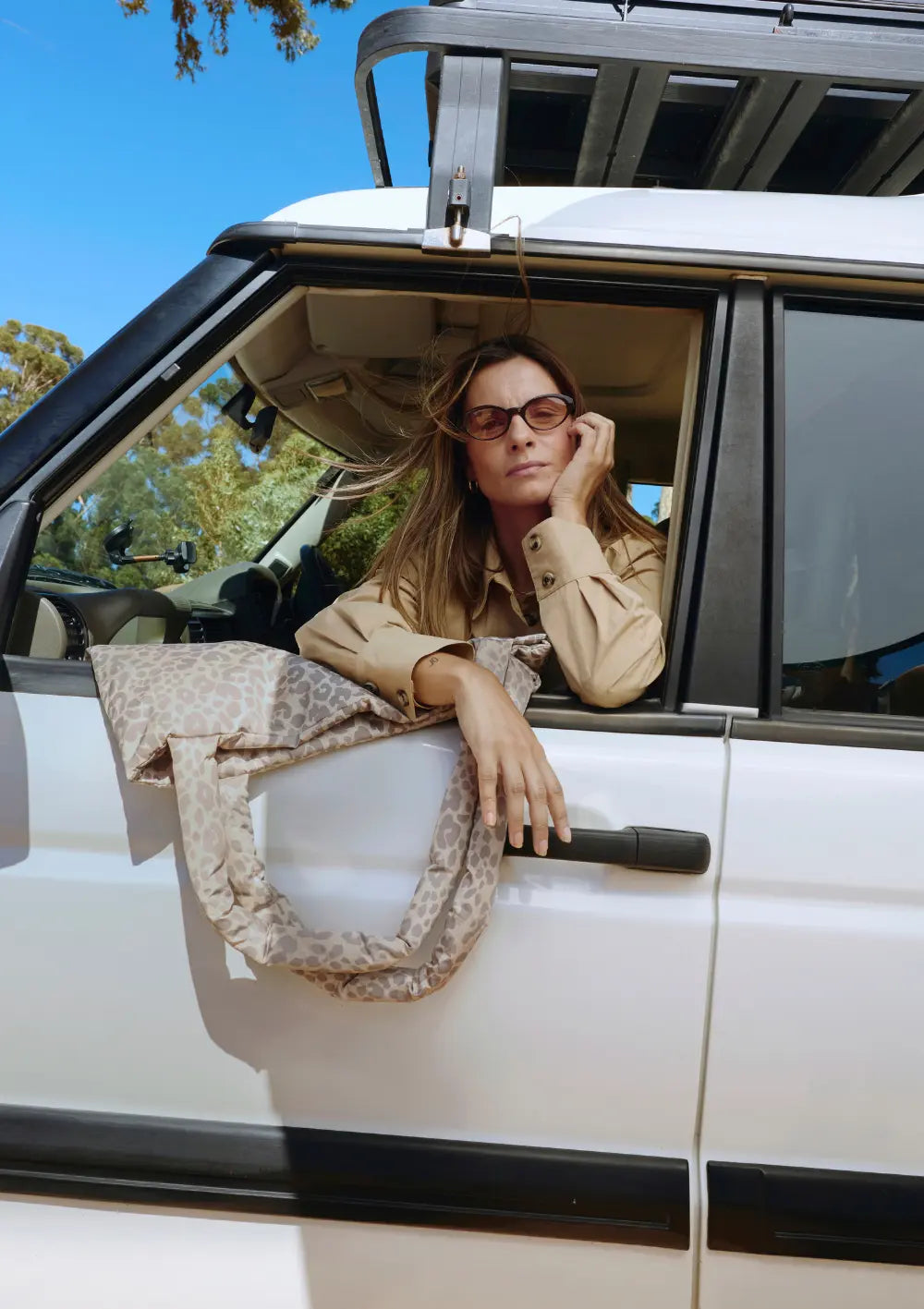 Eine Frau mit Sonnenbrille blickt aus dem Fenster eines weißen Geländewagens, während die weich wattierte Studio Noos Tasche Puffy Mom Bag Beige Leopard lässig über der Autotür hängt.