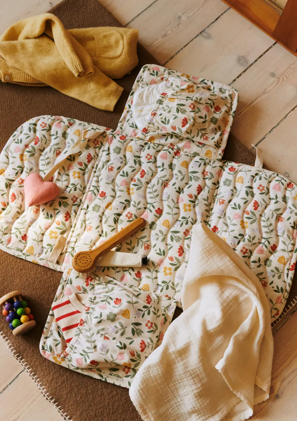 Die kompakt auffaltbare Dear April Wickelunterlage River Flower Market präsentiert sich im bunten Blumen-Design, ausgelegt mit Babyutensilien wie einer Bürste und Kleidung auf einer Decke.