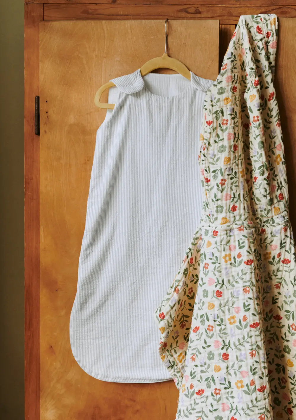 Der ärmellose Dear April Schlafsack Sylvia TOG 1.0 Dew Stripe im feinen Streifen-Design hängt an einem Holzkleiderbügel vor einer hölzernen Schranktür neben einer floral gemusterten Steppdecke.