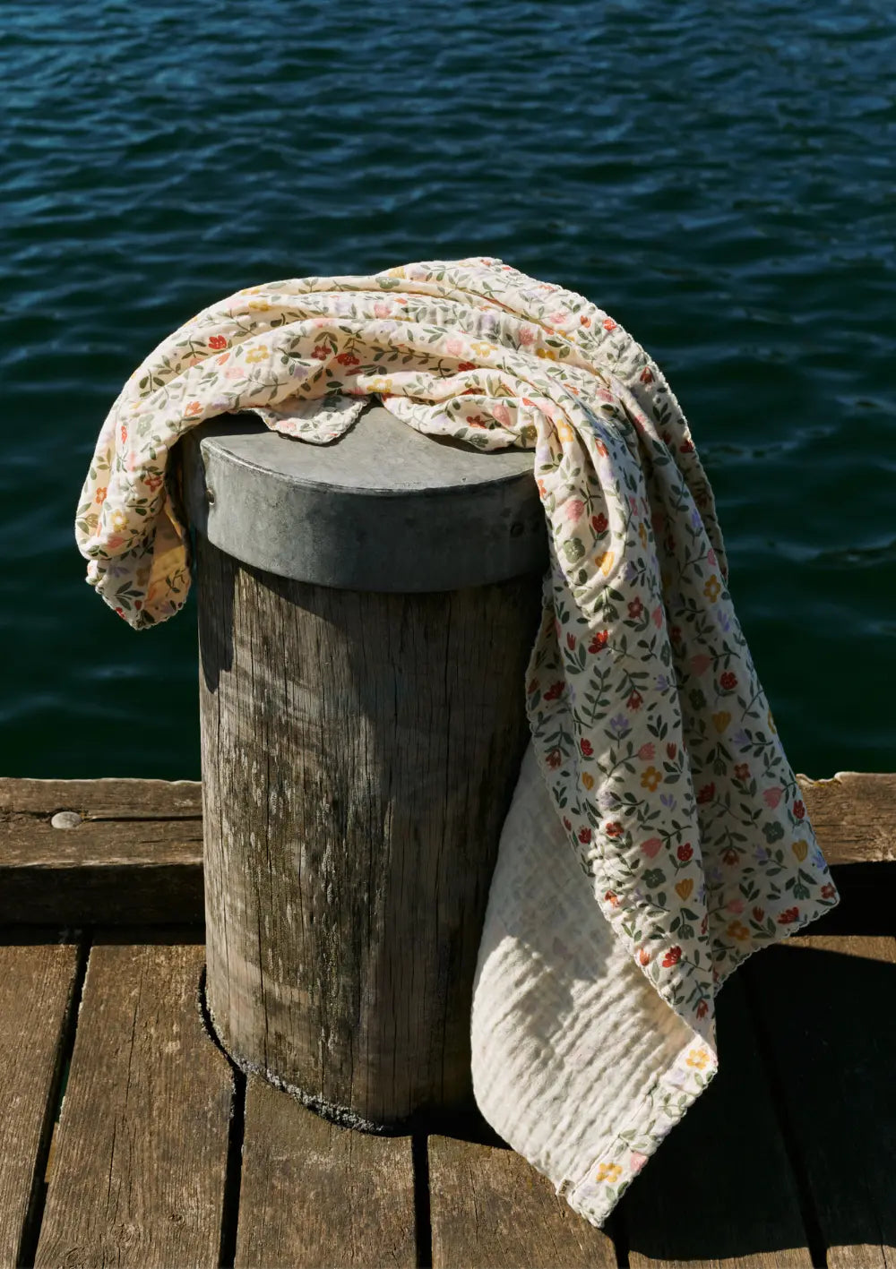 Die fein gemusterte Dear April Musselin Babydecke Murray Flower Market (120 x 120 cm) liegt dekorativ ausgebreitet über einem hölzernen Poller an einer Steg-Kulisse direkt am blauen Wasser.