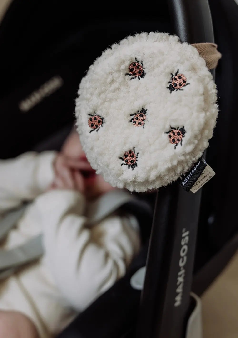 Die runde, weiße Flow Amsterdam Einschlafhilfe für unterwegs Ted Comforter on the go Ladybug aus flauschigem Teddy-Stoff mit aufgestickten Marienkäfern hängt am Tragebügel einer Babyschale, in der ein Baby liegt.