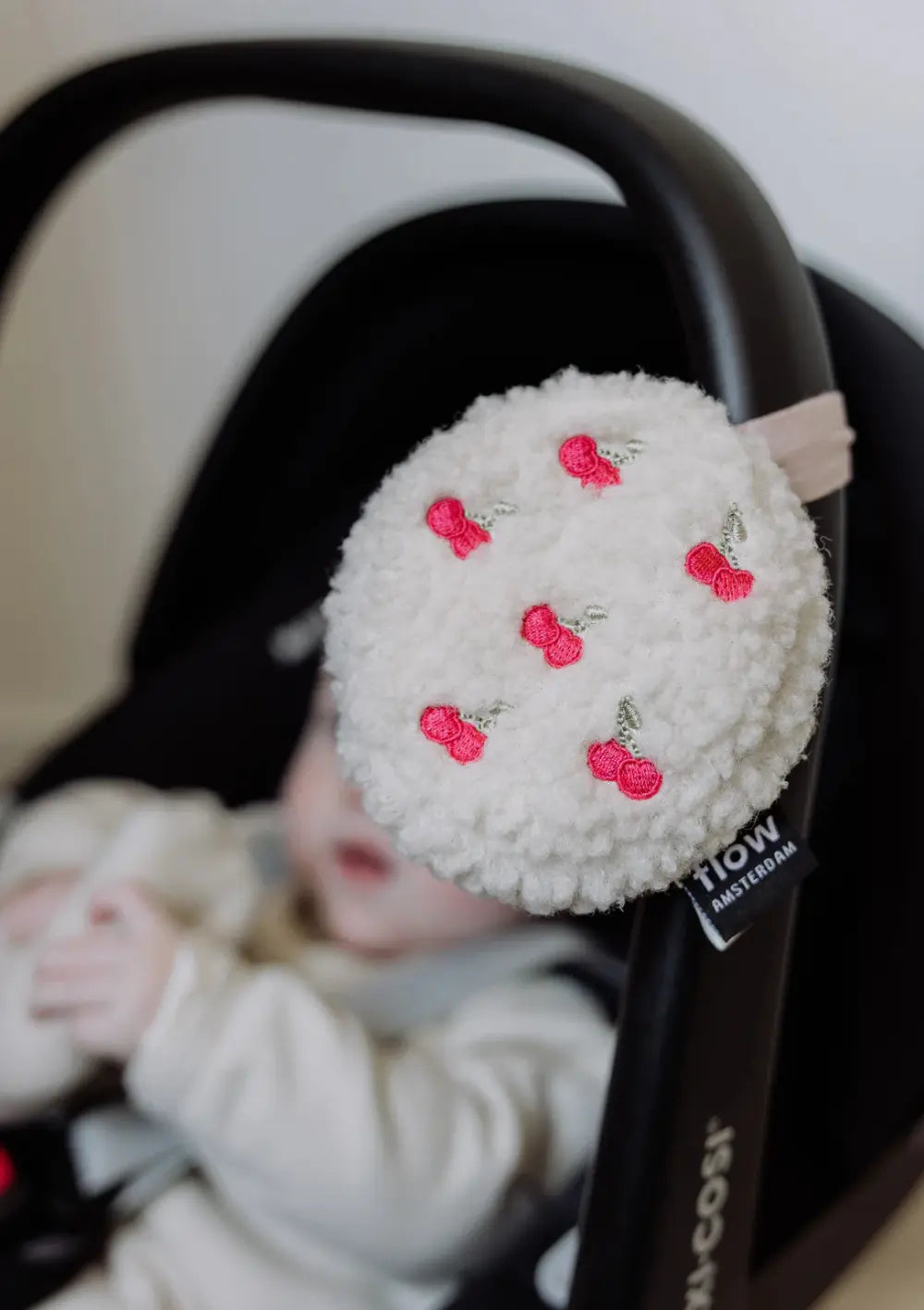 Die runde, weiße Flow Amsterdam Einschlafhilfe für unterwegs Lucy Comforter on the go Cherry aus flauschigem Teddy-Stoff mit aufgestickten Kirschen hängt am Tragebügel einer Babyschale, in der ein Baby liegt.
