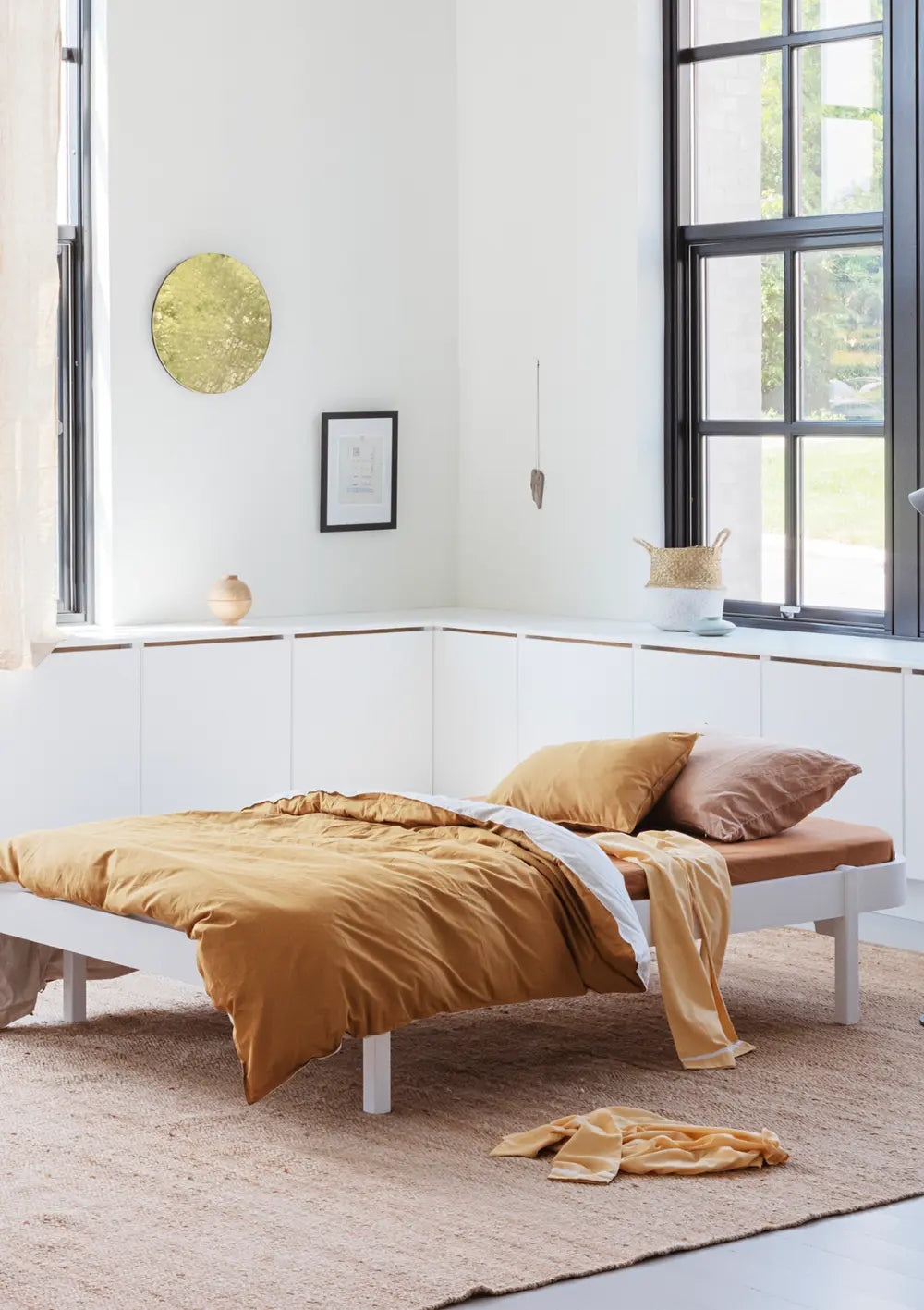 Das minimalistische Oliver Furniture Wood Lounger Bett weiß steht in einem lichtdurchfluteten Raum mit großen Fenstern, bezogen mit einer senfgelben Bettwäsche auf einem großen Naturfaserteppich.