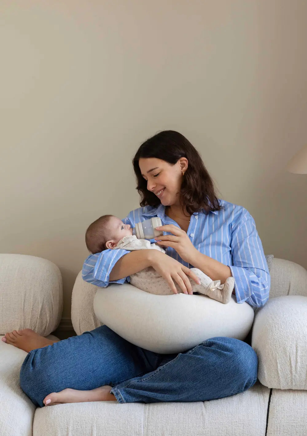 Eine lächelnde Mutter füttert ihr Baby mit der Flasche auf dem Sofa und bettet das Kind dabei komfortabel auf dem u-förmig angepassten bbhugme Stillkissen Soft White.
