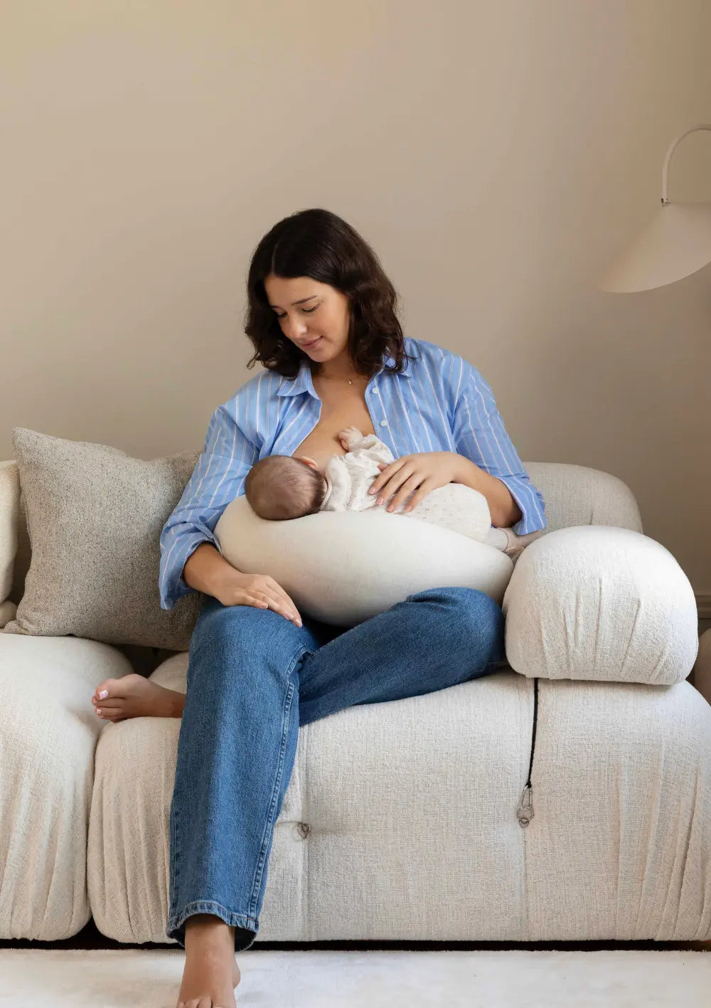 Eine Mutter sitzt im bequemen Schneidersitz auf einer hellen Couch und nutzt das cremeweiße bbhugme Stillkissen Soft White, um ihr Neugeborenes beim Stillen stabil und ergonomisch zu stützen.