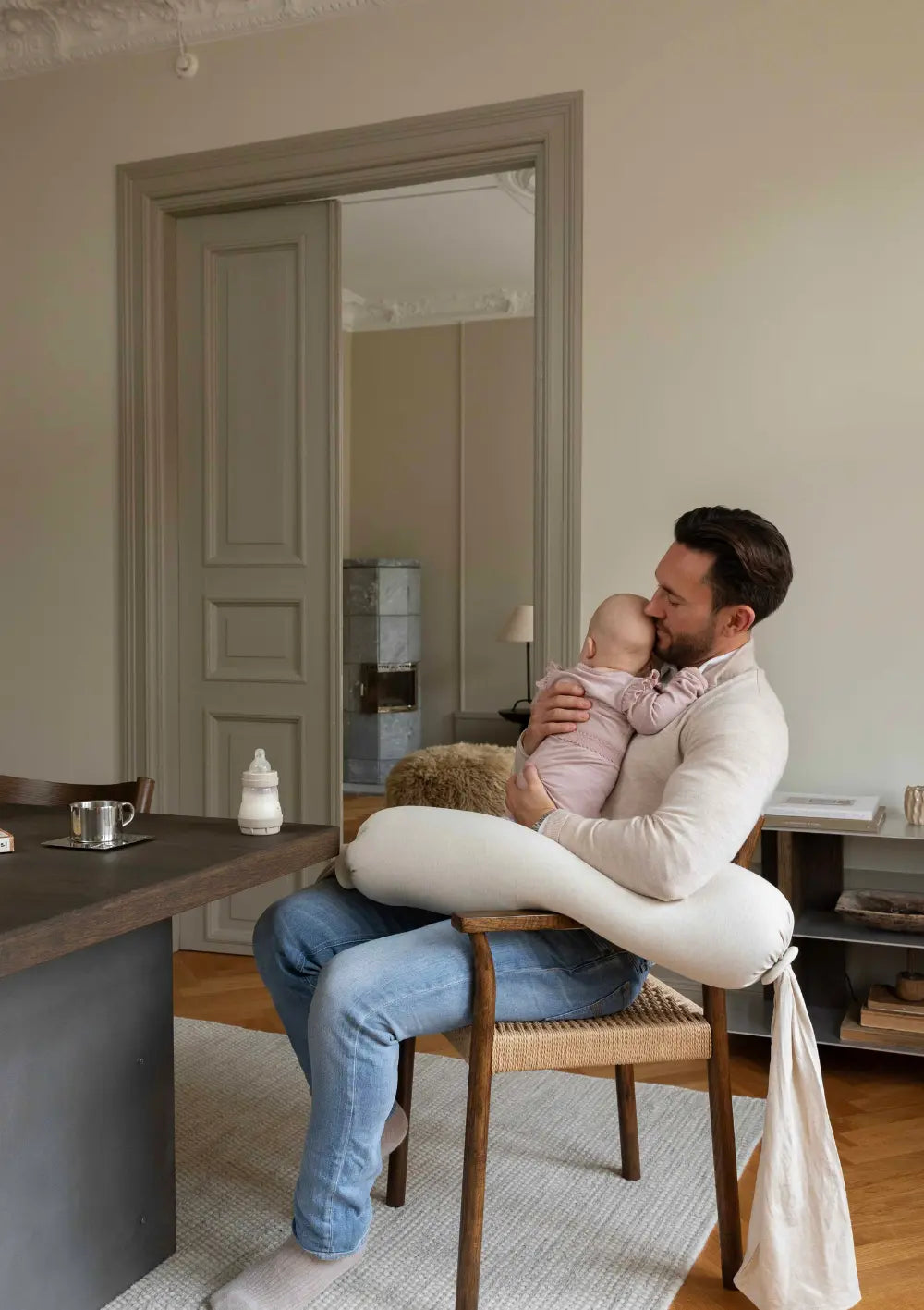 Ein Vater sitzt auf einem Flechtstuhl am Esstisch, hält sein Baby im Arm und nutzt das weiche bbhugme Stillkissen Soft White zur entlastenden Unterlagerung des Säuglings im Alltag.