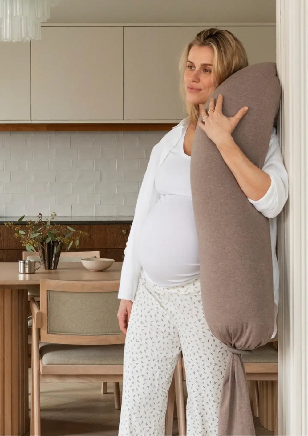 Eine schwangere Frau steht im Wohnbereich an eine Wand gelehnt und hält das große, weiche bbhugme Schwangerschaftskissen Hazel Brown aufrecht im Arm; im Hintergrund ist eine minimalistische Küche erkennbar.