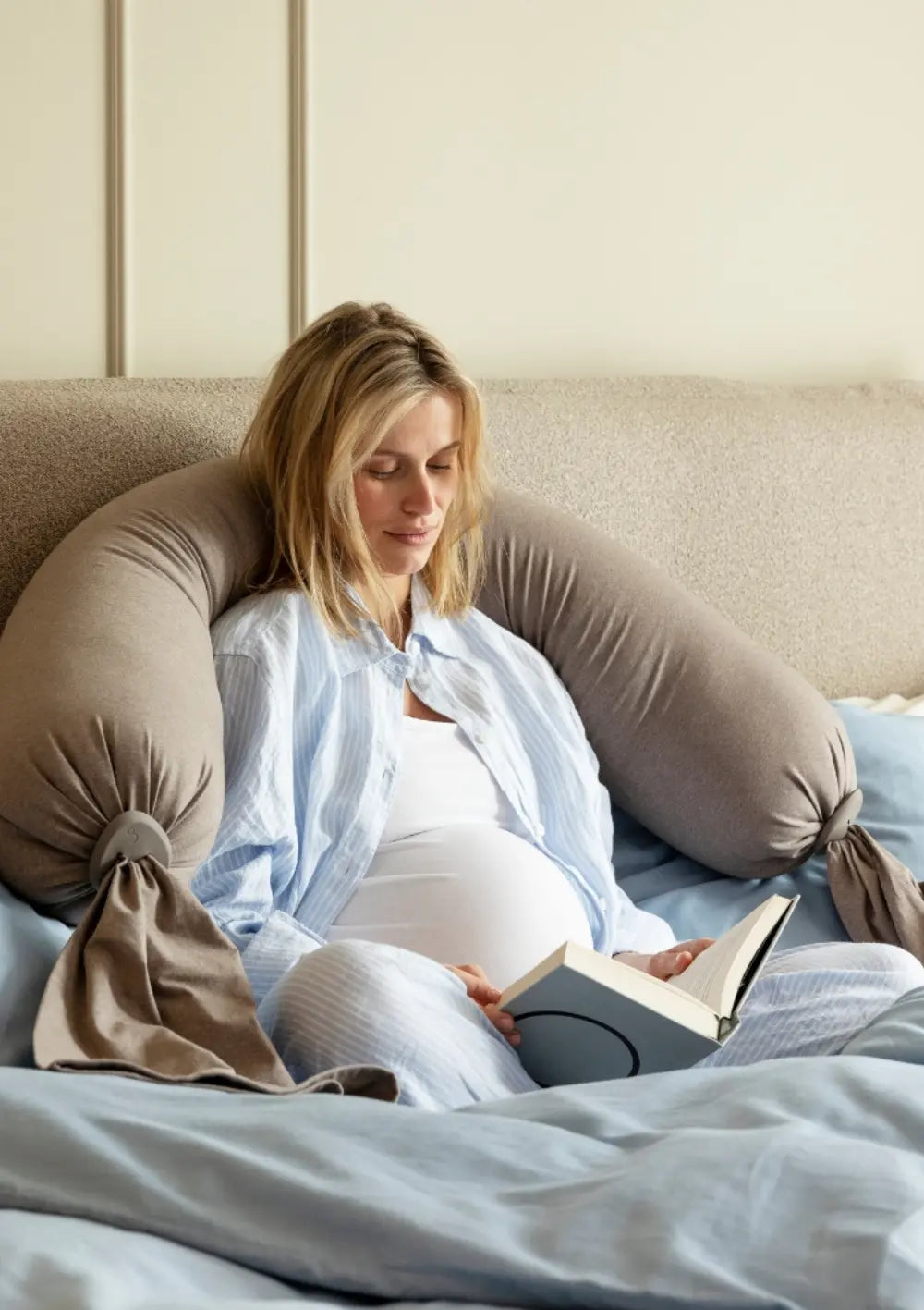 Eine schwangere Frau sitzt aufrecht im Bett und liest ein Buch, während das u-förmig hinter ihr platzierte bbhugme Schwangerschaftskissen Hazel Brown als ergonomische Rücken- und Armlehne dient.