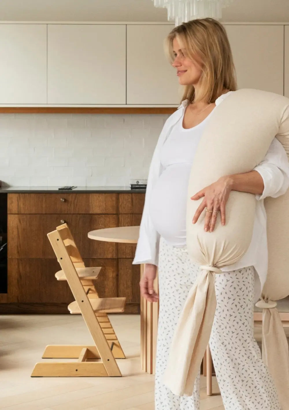 Eine schwangere Frau steht im Wohnbereich und hält das große, weiche bbhugme Schwangerschaftskissen Soft White aufrecht im Arm; im Hintergrund ist eine moderne Küche mit einem Holz-Hochstuhl zu sehen.