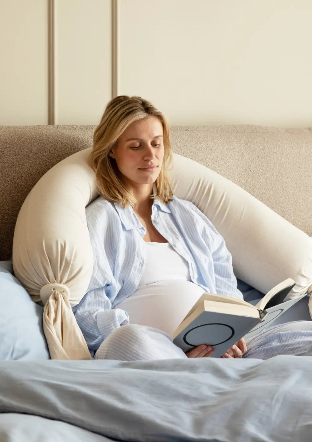 Eine schwangere Frau sitzt aufrecht im Bett und liest ein Buch, während das u-förmig hinter ihr platzierte, cremeweiße bbhugme Schwangerschaftskissen Soft White als ergonomische Rücken- und Armlehne dient.