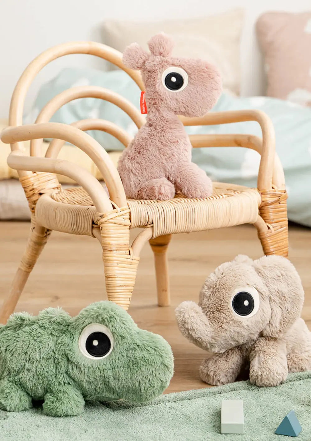 Drei weiche Done by Deer Kuscheltiere im Kinderzimmer: Giraffe Raffi sitzt auf einem kleinen Rattanstuhl, während das grüne Krokodil Croco und der beige Elefant Elphee davor auf dem Boden liegen.