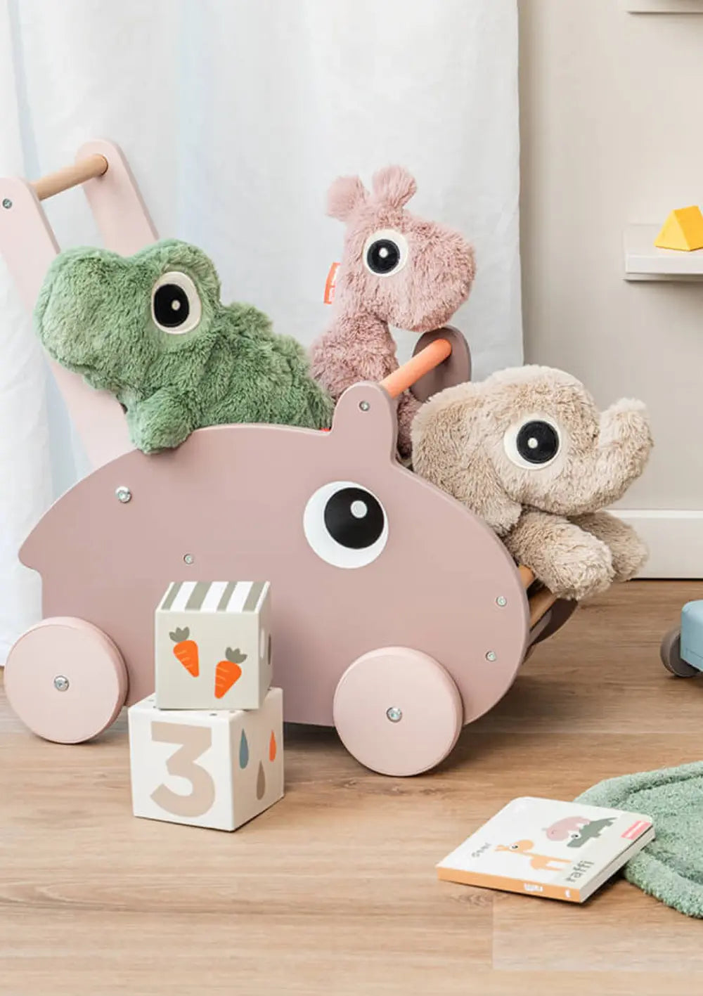 Drei plüschige Done by Deer Kuscheltiere sitzen in einem rosafarbenen Spielzeug-Laufwagen aus Holz, darunter das grüne Krokodil Croco, der beige Elefant Elphee und die altrosa Giraffe Raffi.