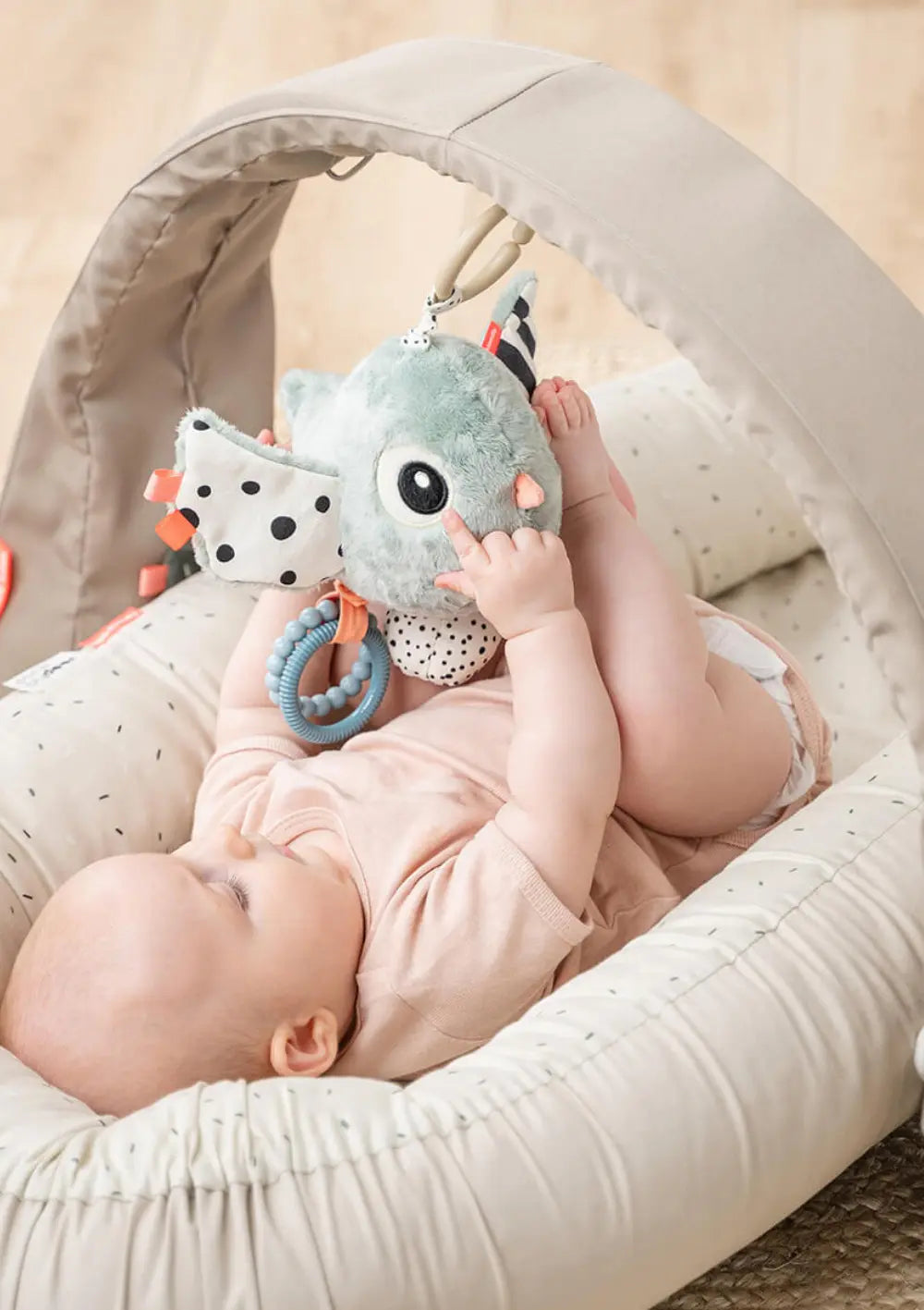 Ein im Nestchen liegendes Baby spielt  mit dem Done by Deer Spielzeuganhänger 'Birdee' blau, der n einem Spielbogen befestigt ist.