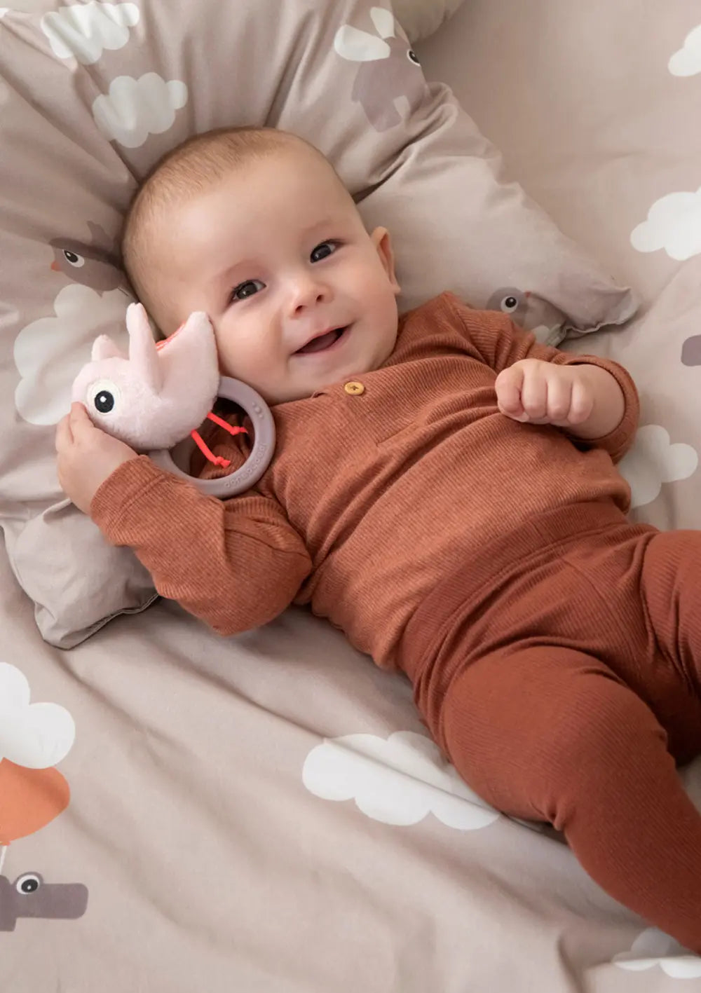 Ein lächelndes Baby liegt im Bett und hält die Done by Deer Rassel mit Beißring 'Birdee' rosa in einer Hand.