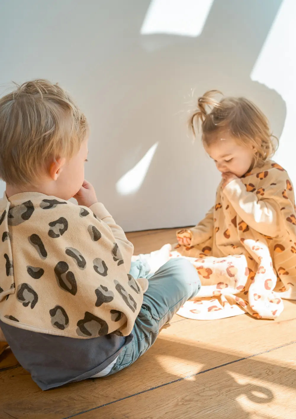Zwei kleine Kinder sitzen auf dem hellen Holzboden und essen einen Snack, optimal geschützt durch das sandfarbene Little Casimir Langarm-Lätzchen Boo Desert Dune mit seinem modernen, dunklen Leoparden-Muster.
