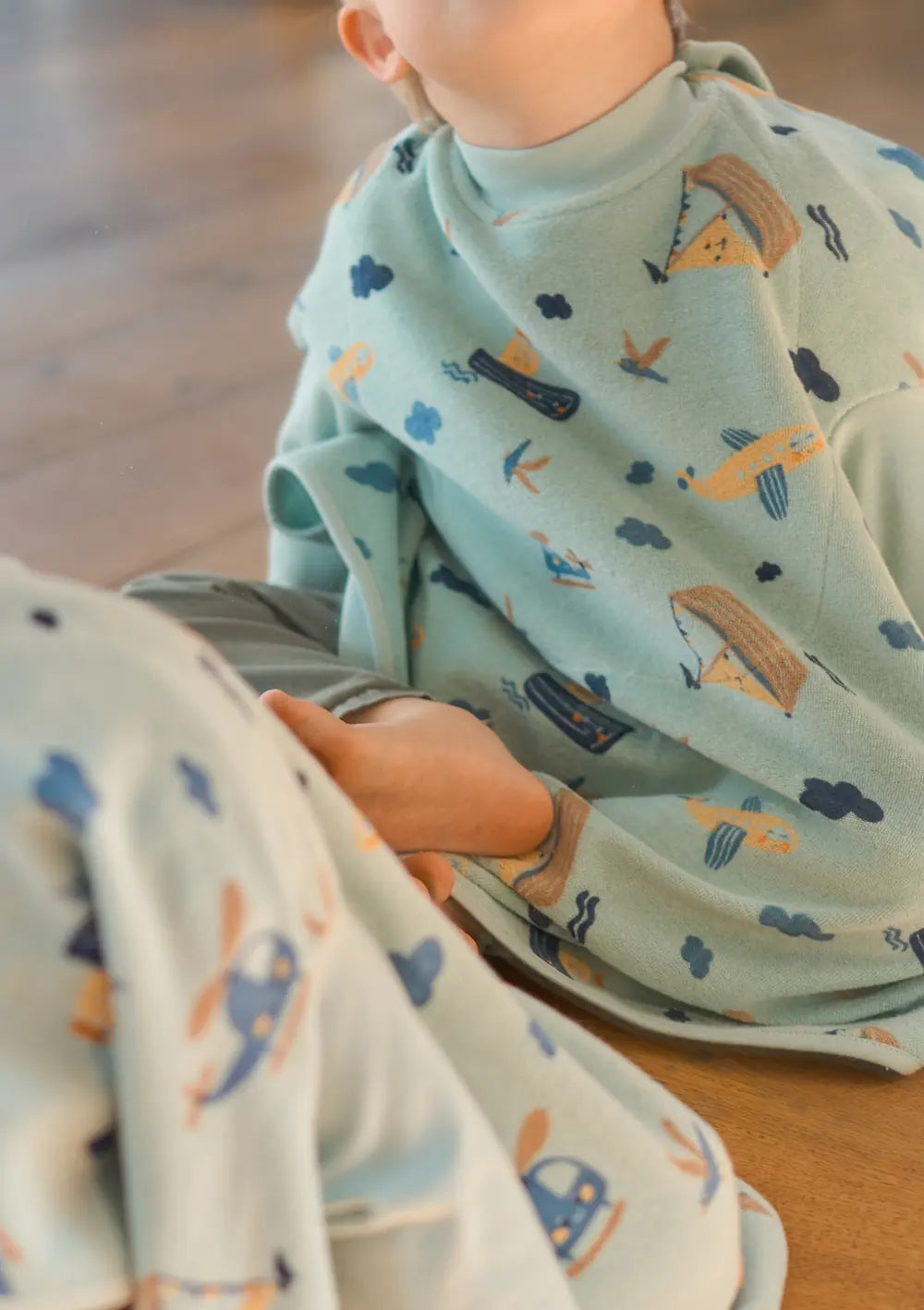 Nahaufnahme eines Kindes, das im hellblauen Little Casimir Langarm-Lätzchen Boo Little Voyagers auf dem Boden sitzt, während sich das verspielte Muster aus kleinen Booten, Hubschraubern und Wolken spiegelt.