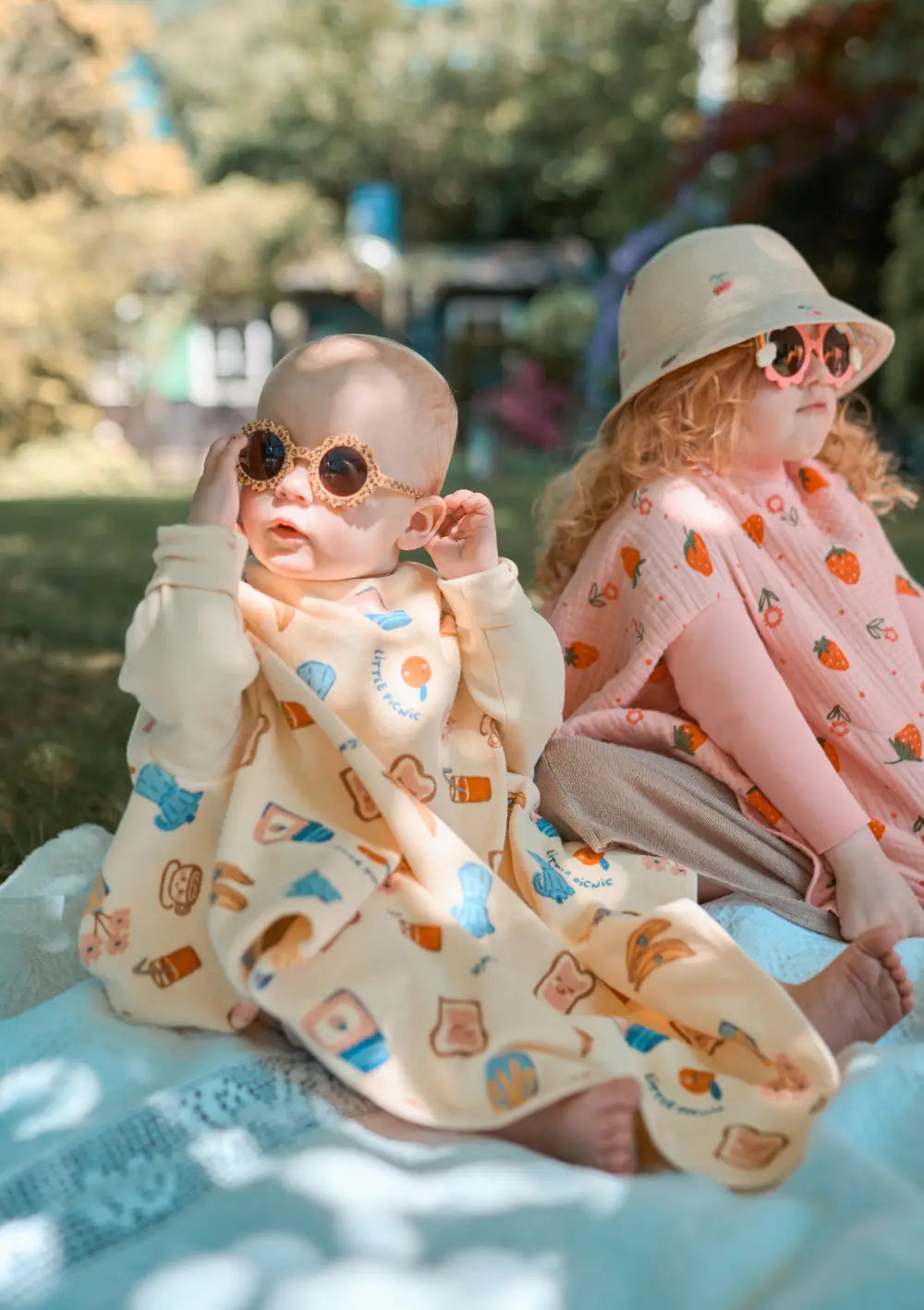 Zwei Kinder sitzen an einem sonnigen Tag im Freien auf einer Decke; das vordere Kleinkind trägt das hellgelbe Little Casimir Langarm-Lätzchen Boo Picnic Club mit buntem Print und rückt sich eine Sonnenbrille zurecht.