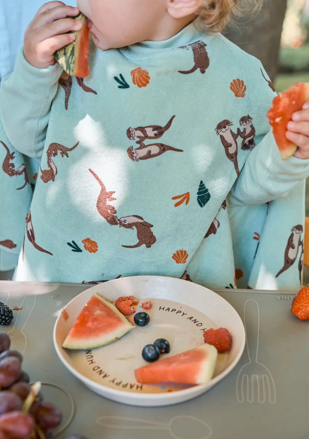 Ein Kleinkind trägt das Little Casimir Langarm-Lätzchen Boo Otter mit verspieltem Otter- und Muschelprint und isst am Tisch frische Wassermelone von einem Kinderteller.