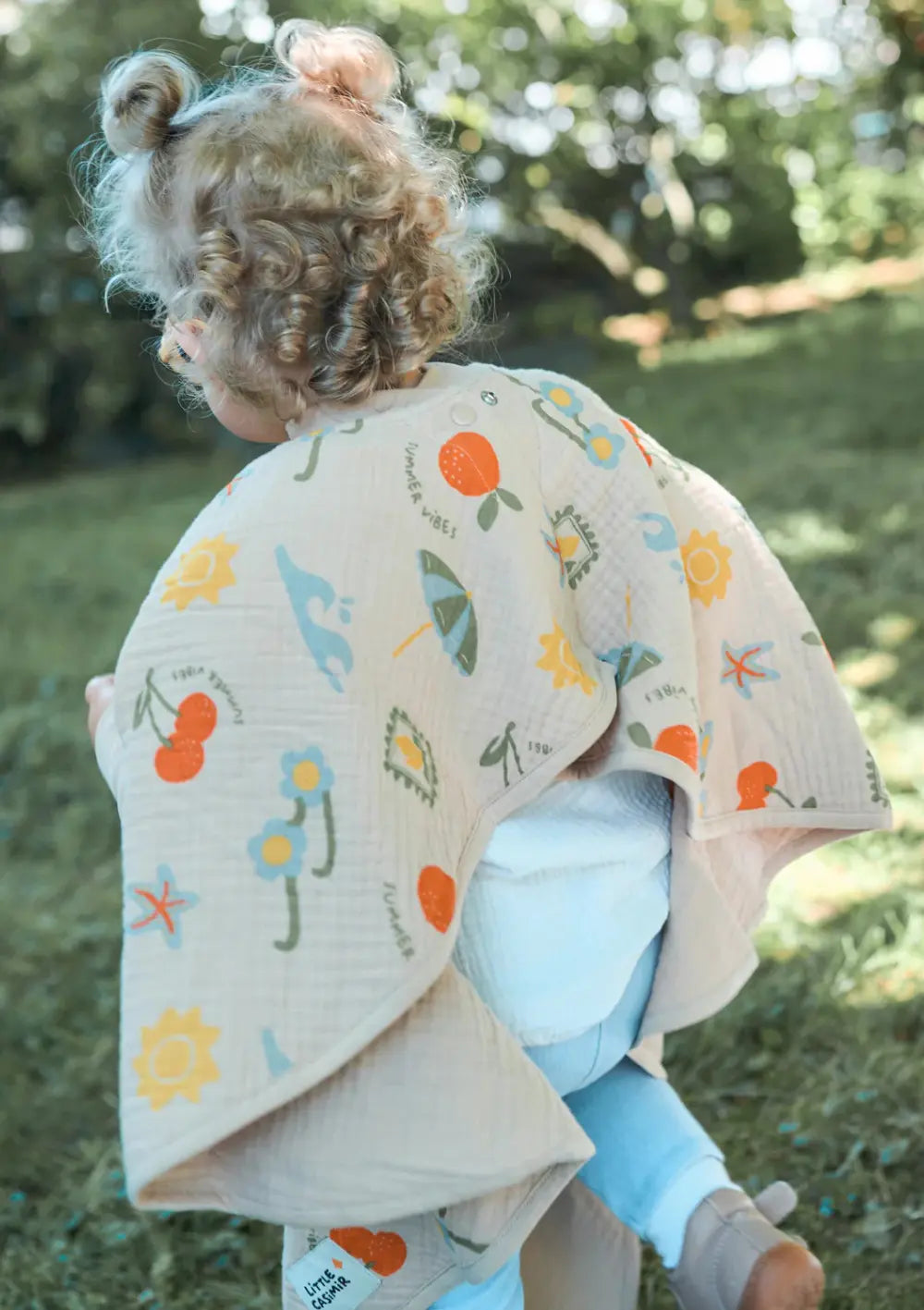 Rückansicht eines kleinen Kindes mit Locken, das im Garten über den Rasen läuft; es trägt das luftige, beigefarbene Little Casimir Langarm-Lätzchen Boo Breeze Sunshine Gardens aus Musselin mit bunten Sommermotiven wie Sonnen, Kirschen und Sonnenschirmen.