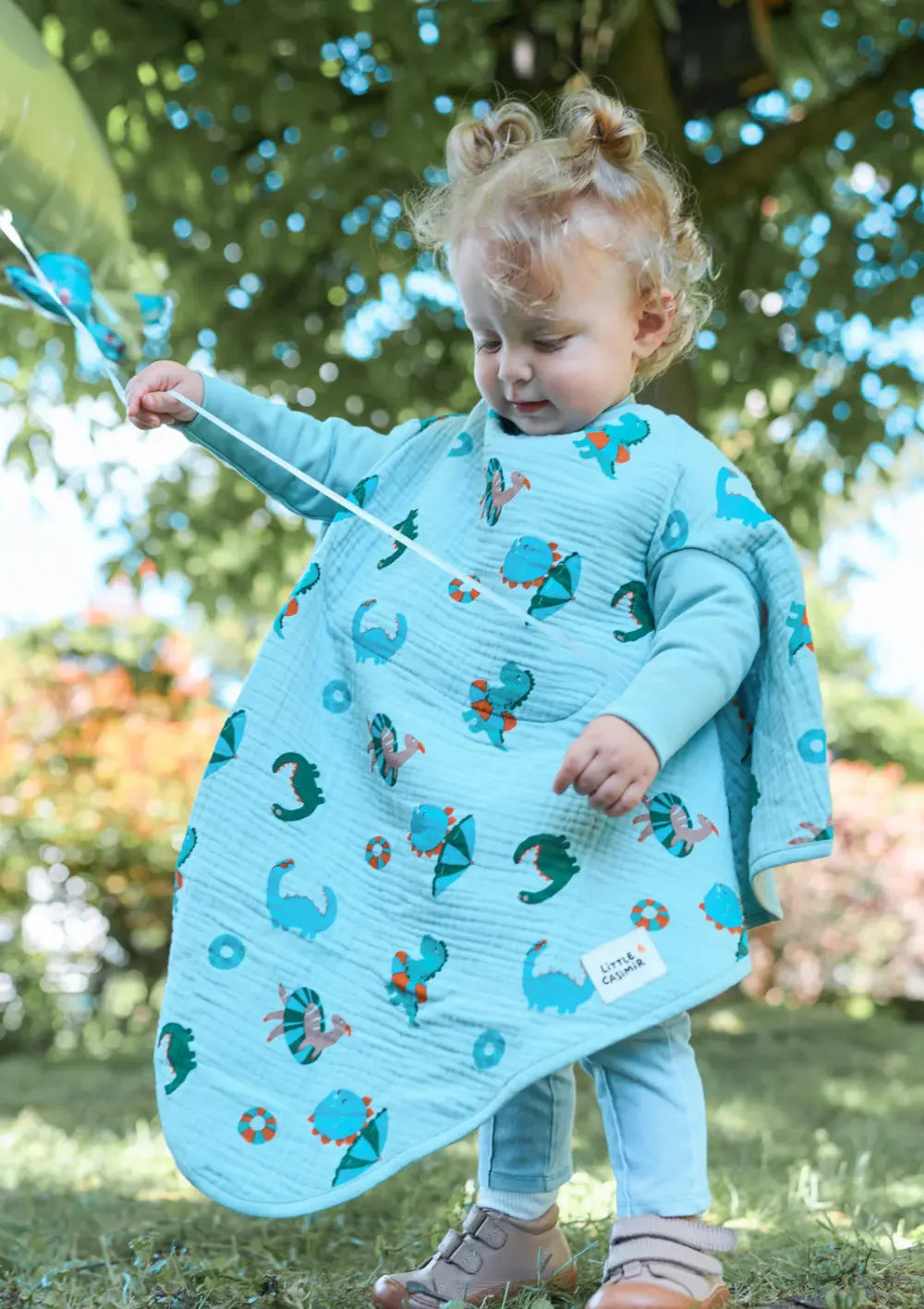 Ein kleines Mädchen spielt im Park. Sie hält die Schnur eines unsichtbaren Ballons in der Hand und schaut nach unten. Sie trägt Little Casimir 'Boo' Breeze Dino Beach Party.