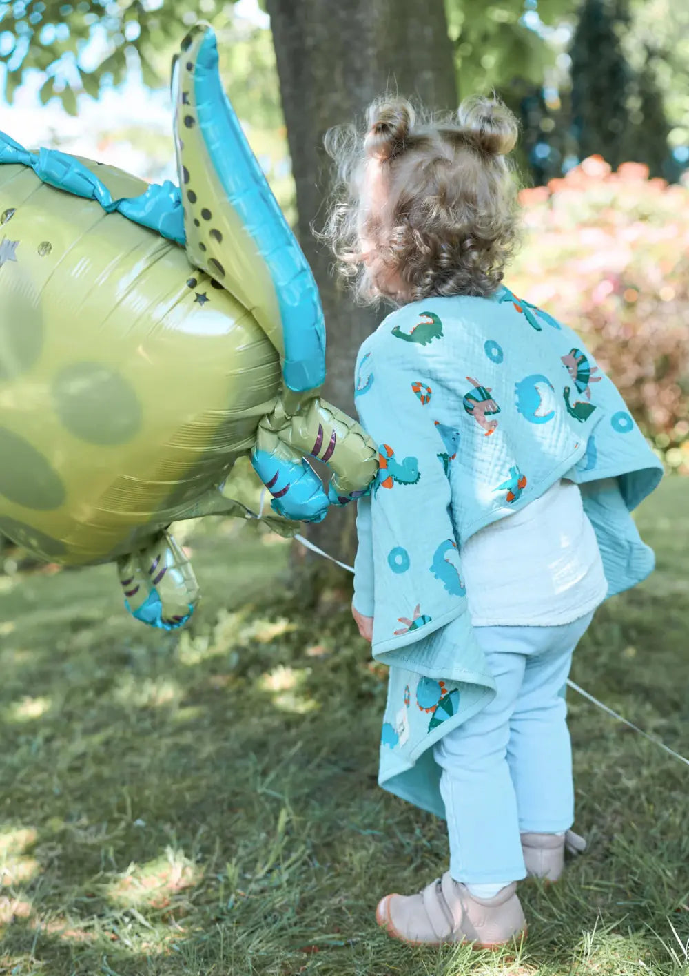 Die Rückansicht eines kleinen Mädchens mit lockigen Haaren, das im Freien auf Gras steht. Sie blickt von der Kamera weg und hält einen großen, grünen und blauen Dinosaurier-Folienballon an einer Schnur in der Hand. Sie trägt das Little Casimir 'Boo' Breeze Dino Beach Party .