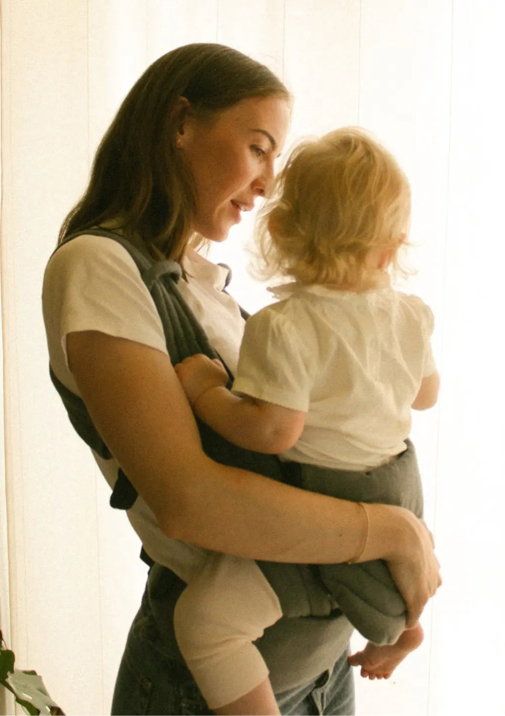 Mutter und Kind stehen an einem Fenster, das Kind sitzt in der Moova Babytrage 'Balance' Atlantic Herringbone