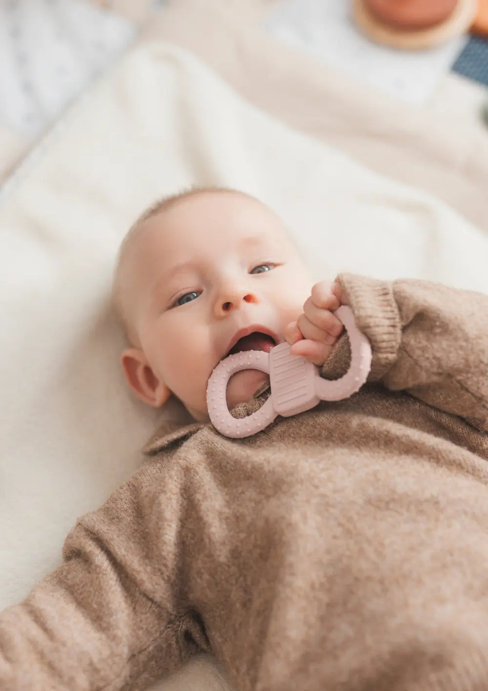 mimiloo Silikon Lern- und Beißring Rose – Baby beißt auf Beißring beim Liegen auf heller Decke.